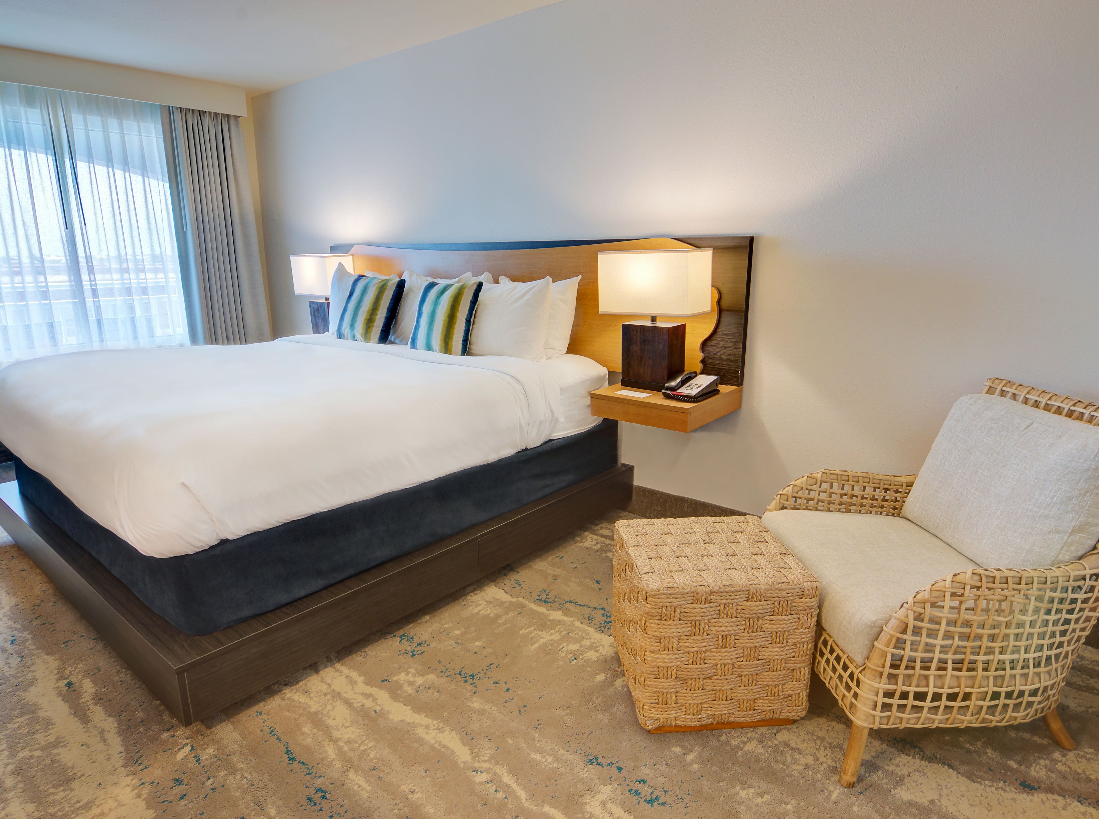 Une chambre d’hôtel avec un grand lit, une table de chevet avec une lampe, un téléphone et un fauteuil en osier.