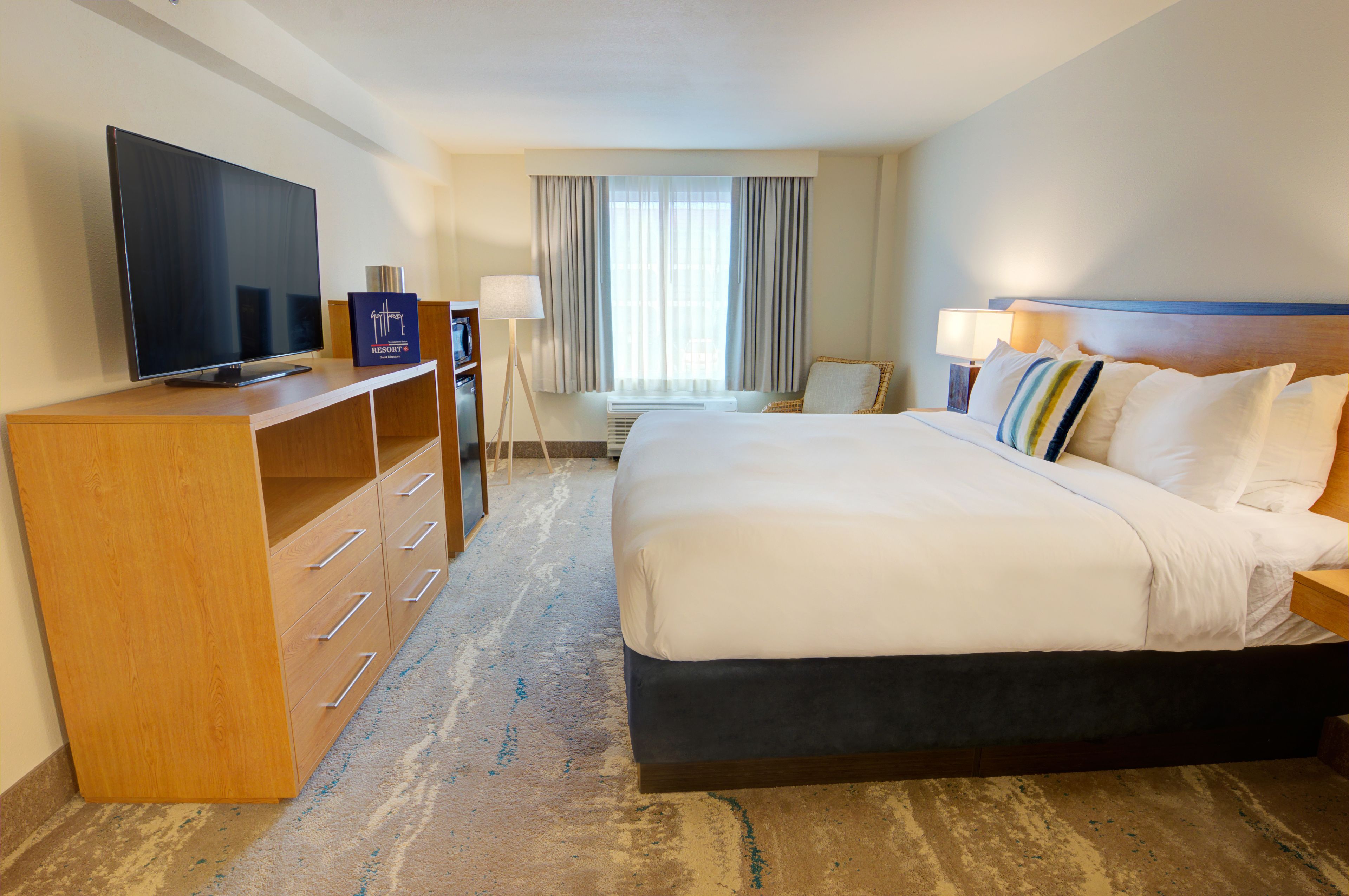 A hotel room with a large bed, a television on a wooden cabinet, a desk with a lamp, and a window with curtains.