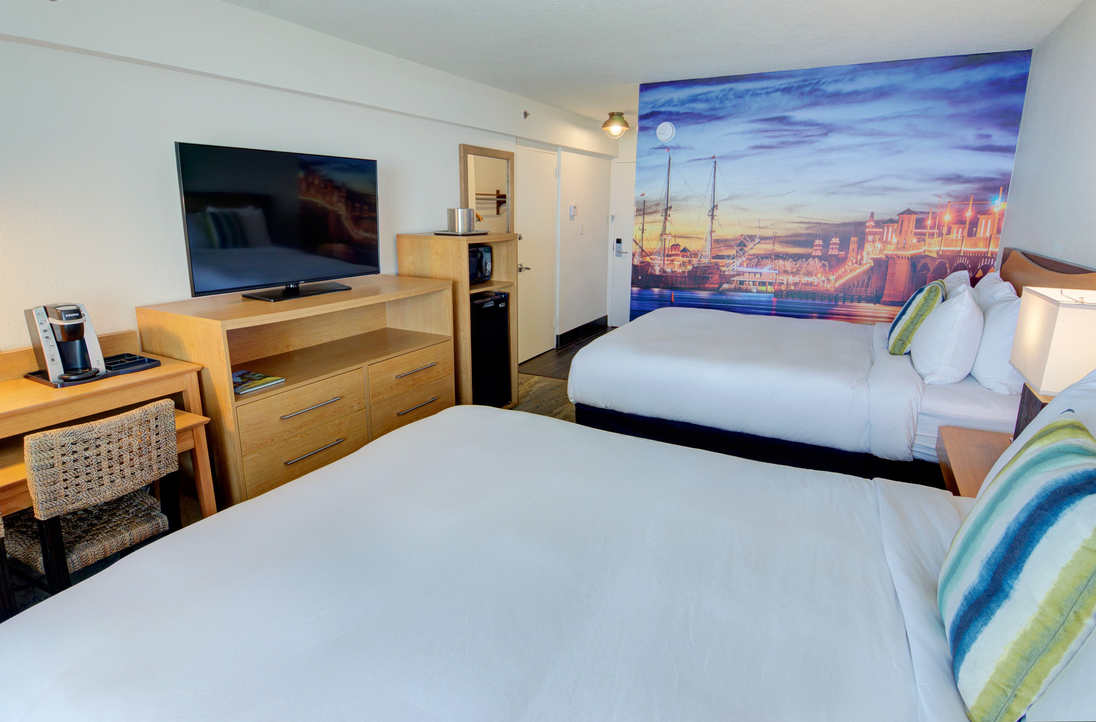 A hotel room with a bed, a desk with a television, a bookshelf, and a painting of a cityscape.