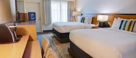 A hotel room with two beds, a desk, a mirror, a lamp, and a view of the outside through the window.