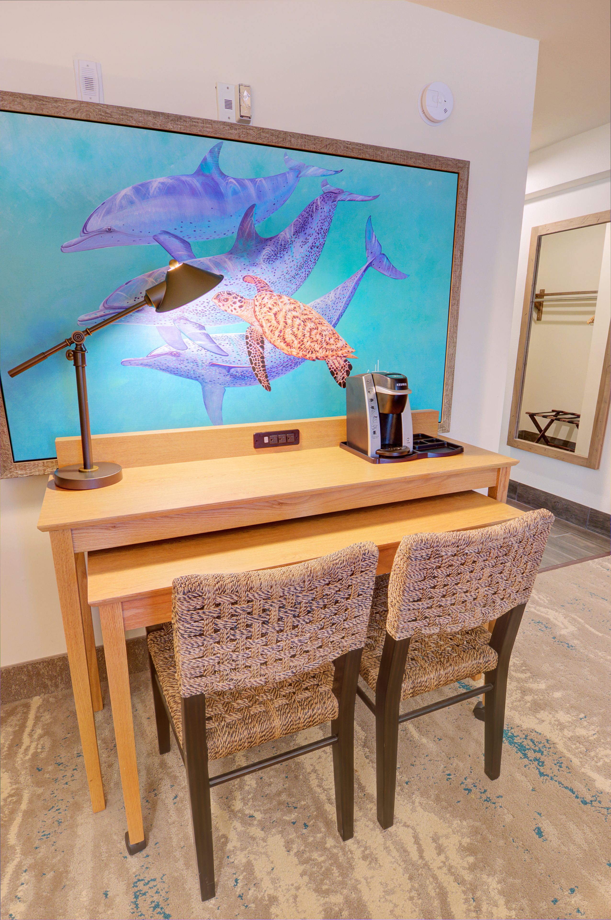 A wooden desk with a coffee maker, two wicker chairs, and a large wall-mounted TV displaying dolphins.