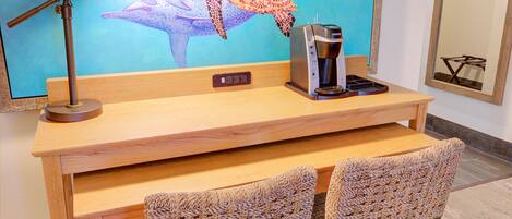 A wooden desk with a coffee maker, two wicker chairs, and a large wall-mounted TV displaying dolphins.