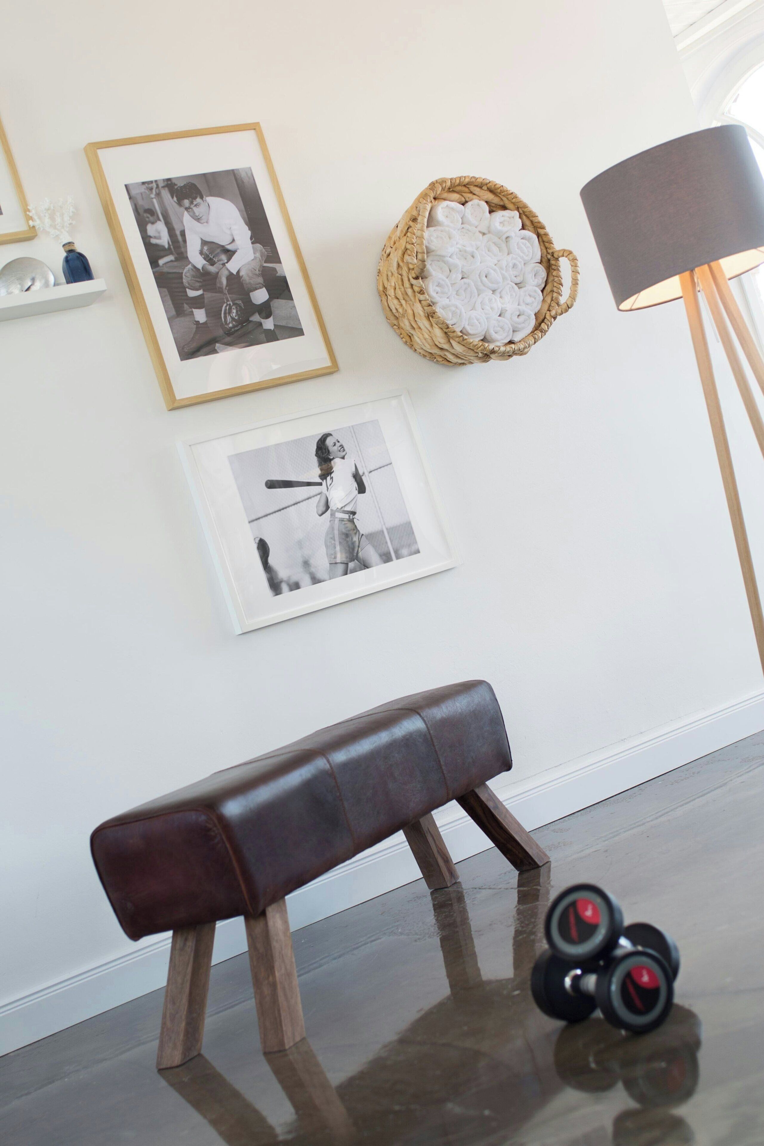 A room with a dark brown bench, framed black and white photographs on the wall, a woven basket with rolled towels, and a floor lamp.