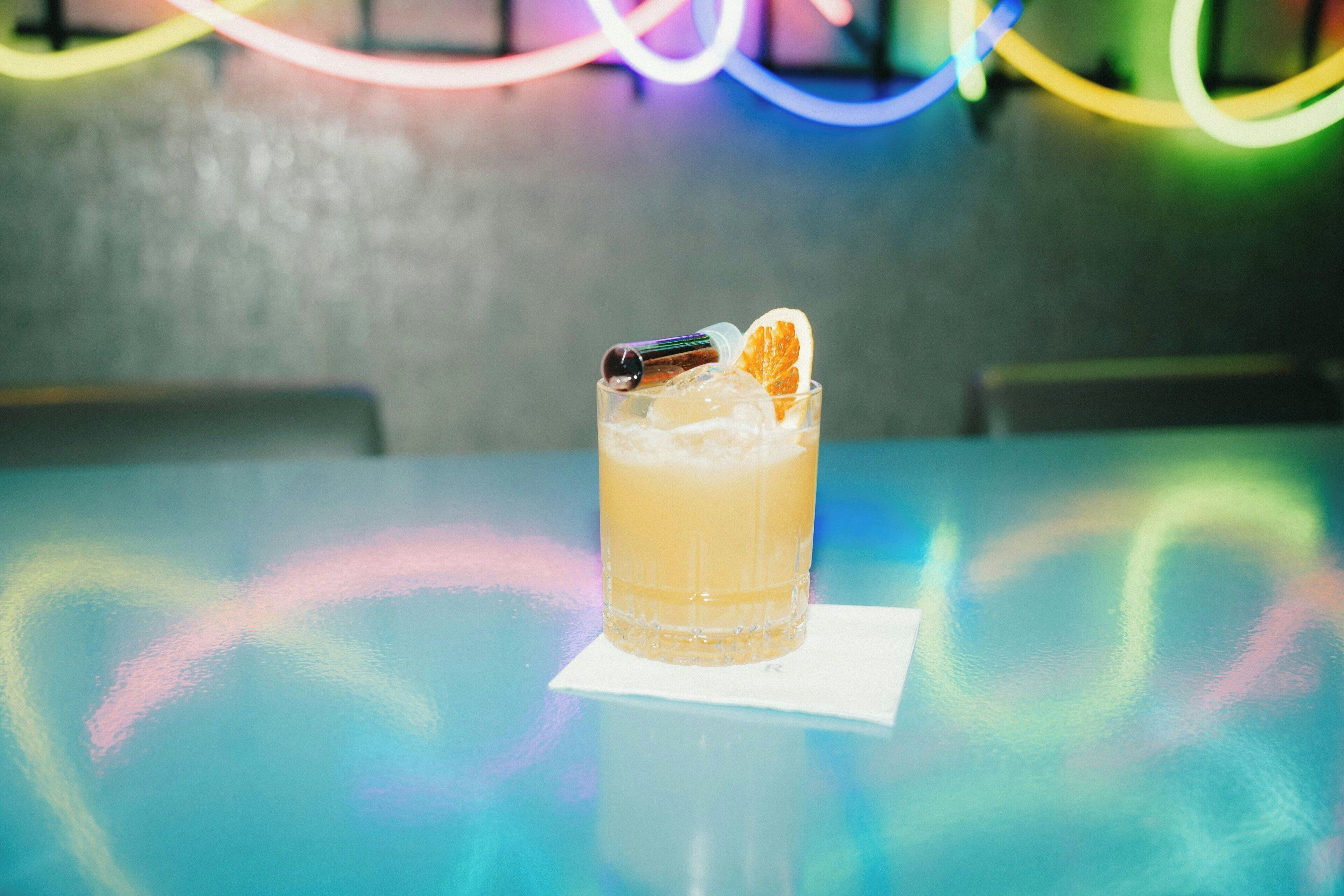 Un vaso de una bebida clara con hielo y una rodaja de naranja, colocado sobre una mesa con superficie reflectante.