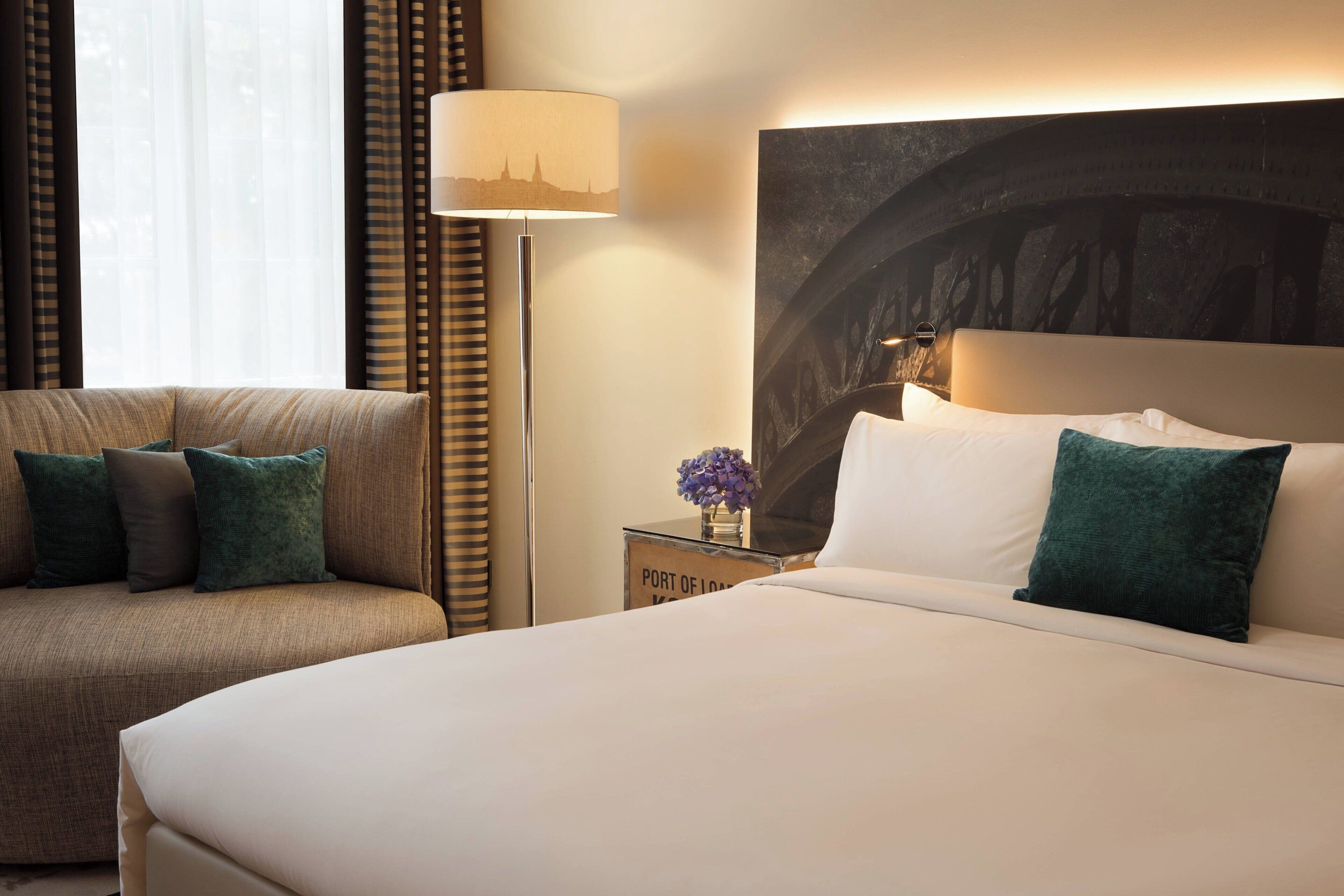 A hotel room with a bed, a sofa, a nightstand with a lamp, and a large wall mural.