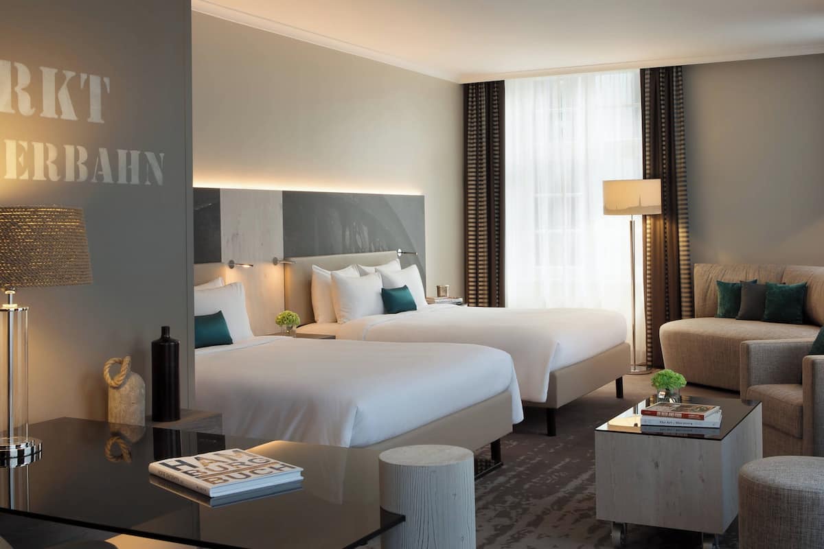 Une chambre d’hôtel avec deux lits, un coin salon avec un canapé et des fauteuils, une petite table avec des livres et une lampe.
