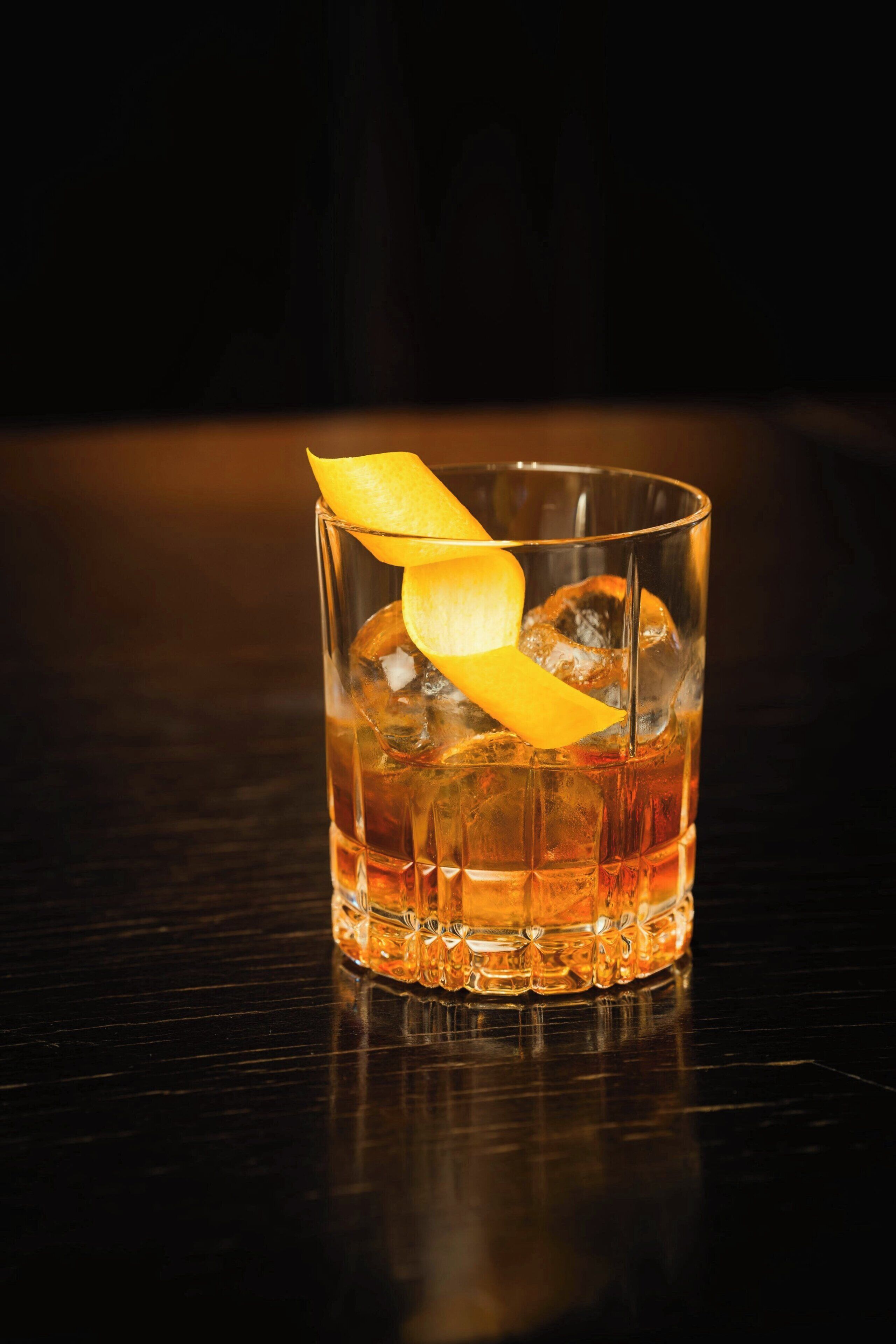 A glass of amber-colored beverage with ice cubes and a lemon peel garnish on a dark surface.