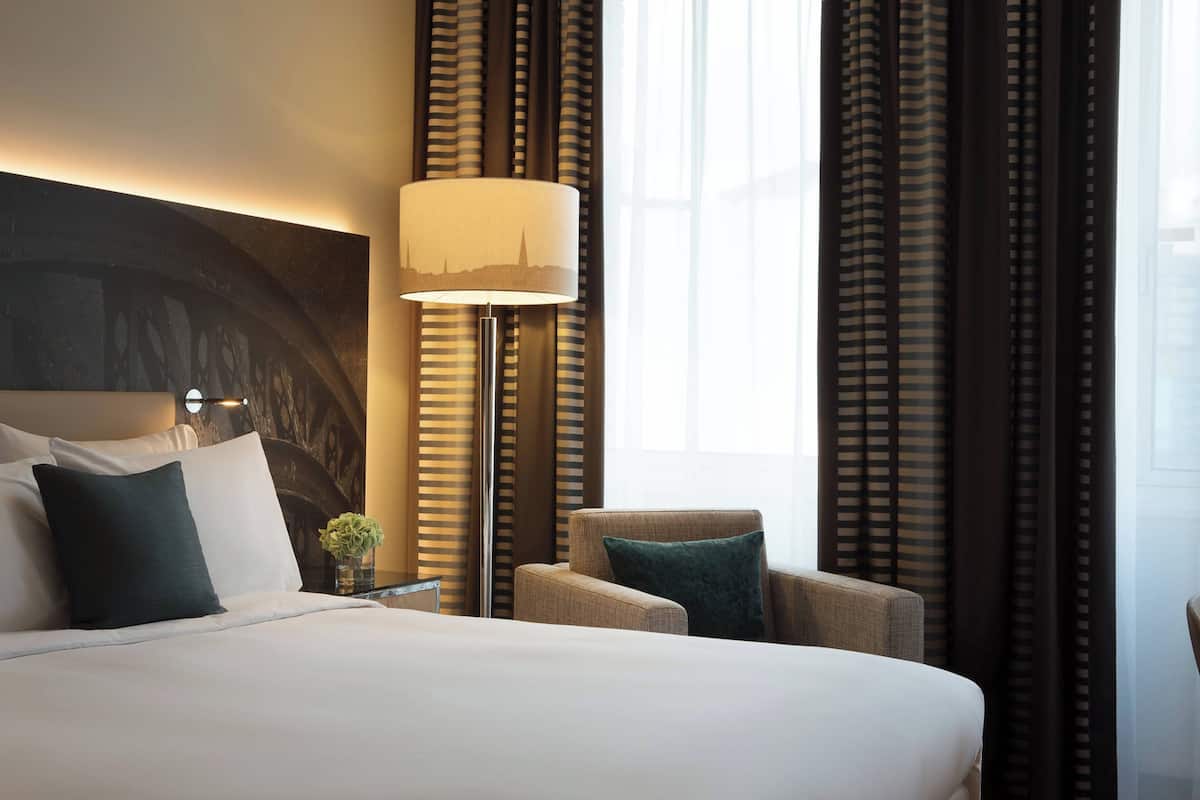 Une chambre d’hôtel avec un grand lit, une table de chevet, une lampe, une chaise et des rideaux.