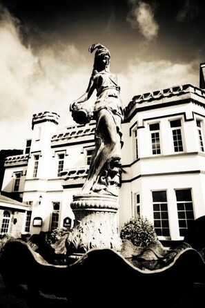 A statue of a woman in a dress and hat, standing on a pedestal in front of a building with a white facade and multiple windows. - Stradey Park Hotel (Llanelli)