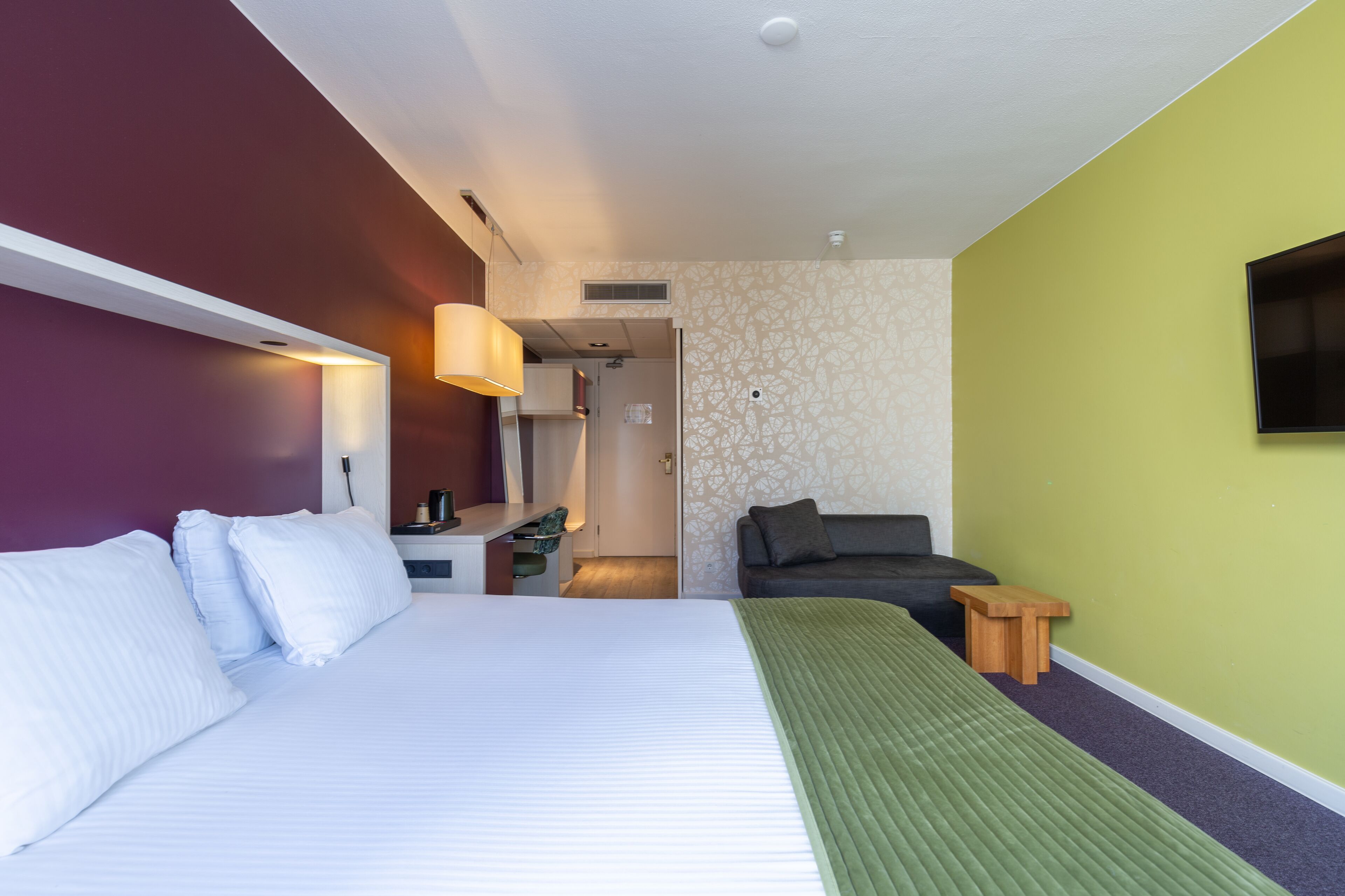A hotel room with a bed, a desk, a chair, a TV, and a small wooden stool.