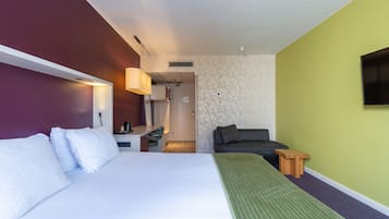 A hotel room with a bed, a desk, a chair, a TV, and a small wooden stool.