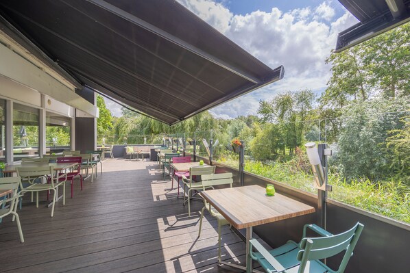 An outdoor patio with wooden tables and chairs, a covered area, and a view of greenery. - Amrath Airport Hotel Rotterdam (Rotterdam)
