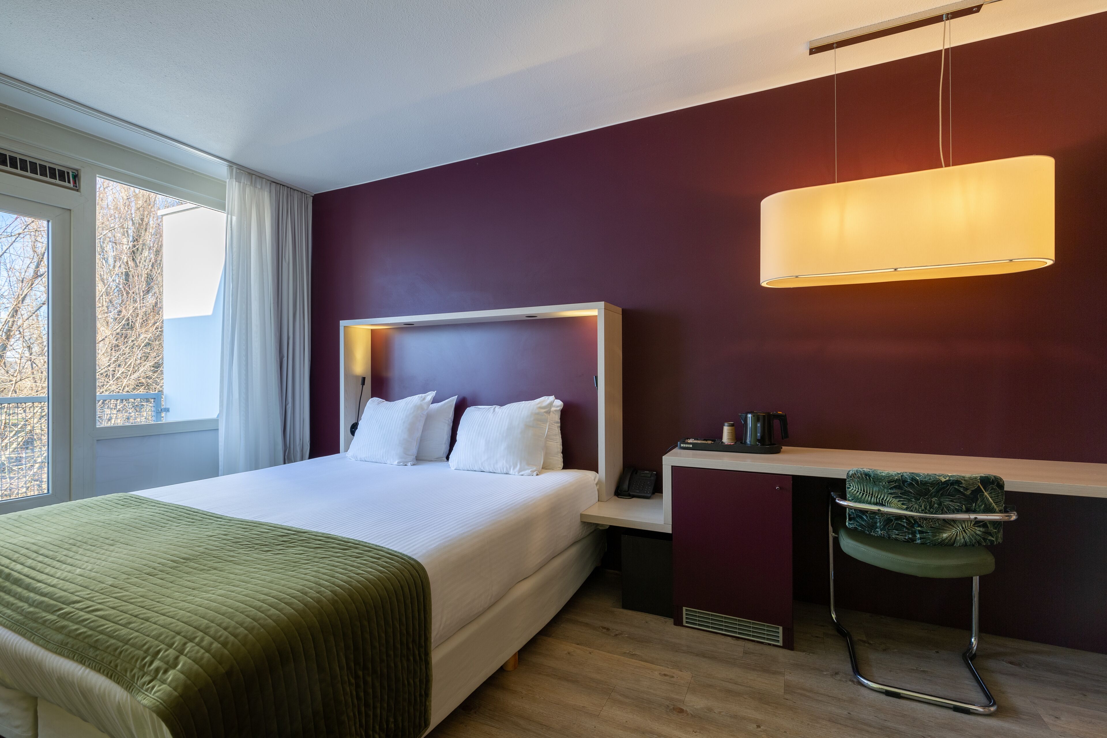 Una habitación de hotel moderna con una cama grande, un escritorio, una silla y un ventanal con cortinas.