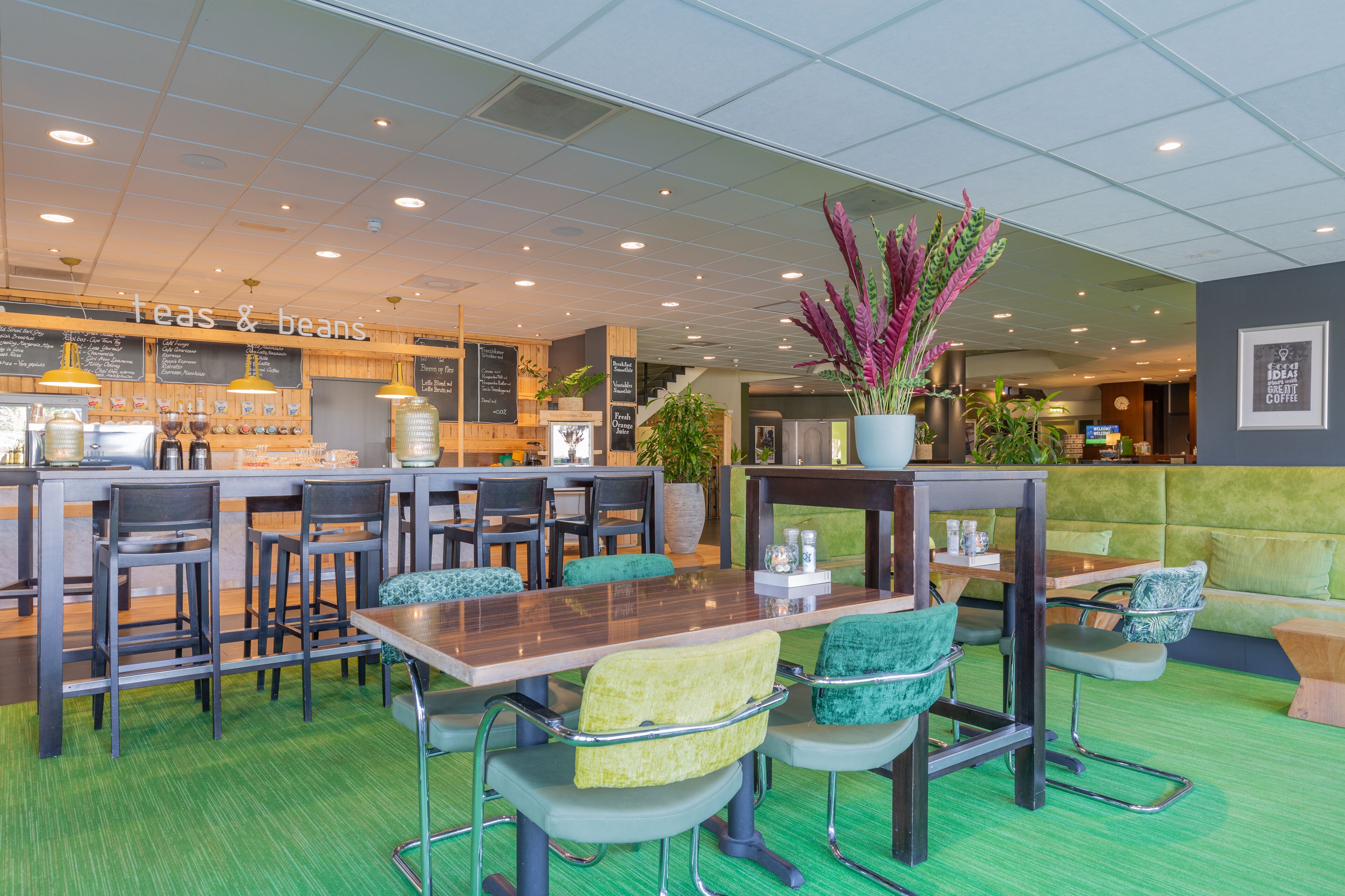 Un restaurante con alfombra verde, mesas y sillas de madera. Hay un letrero que dice "teas & beans" y una planta grande en maceta.