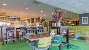 Un restaurante con alfombra verde, mesas y sillas de madera. Hay un letrero que dice "teas & beans" y una planta grande en maceta.