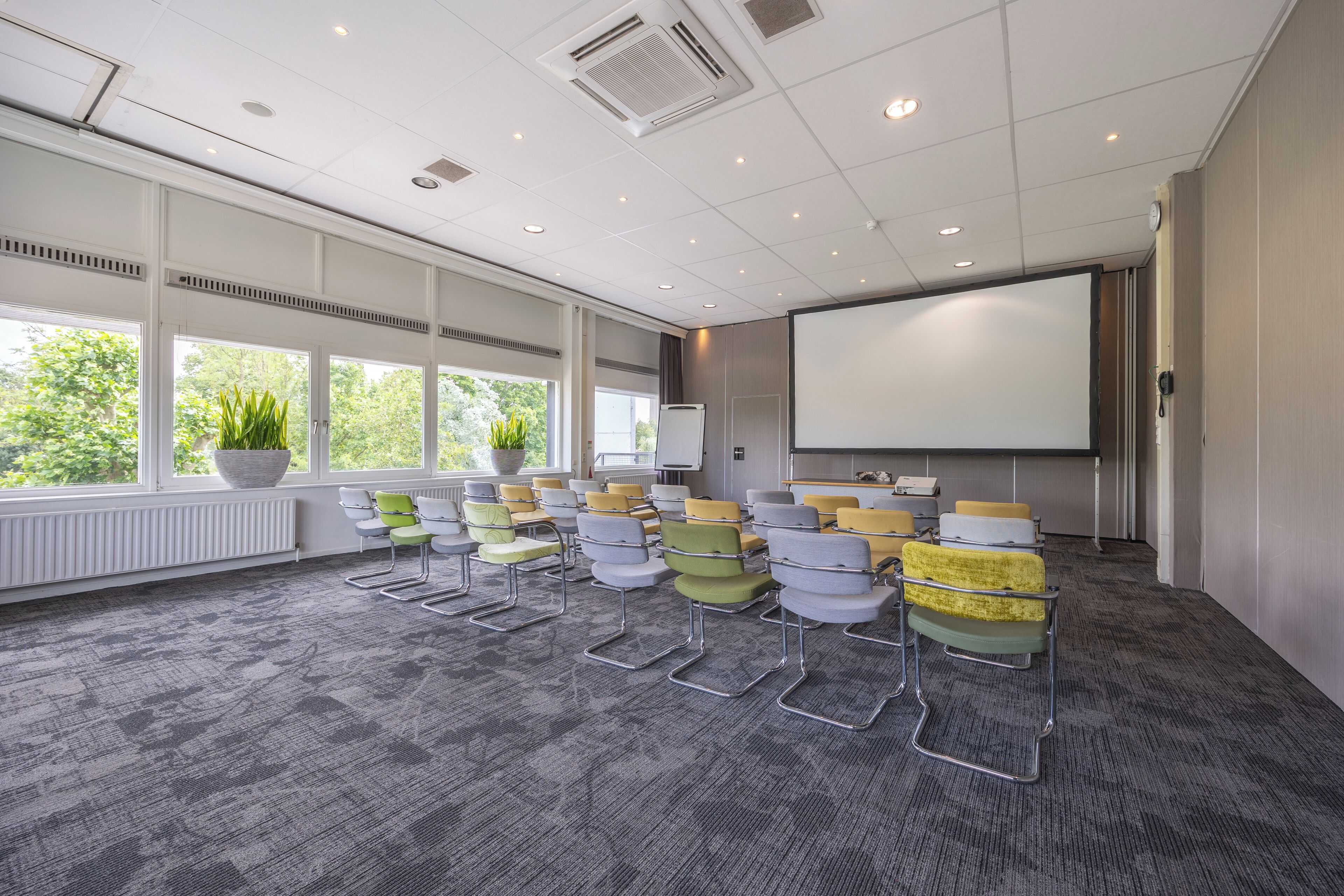 A conference room with rows of chairs, a projector screen, and large windows with plants.