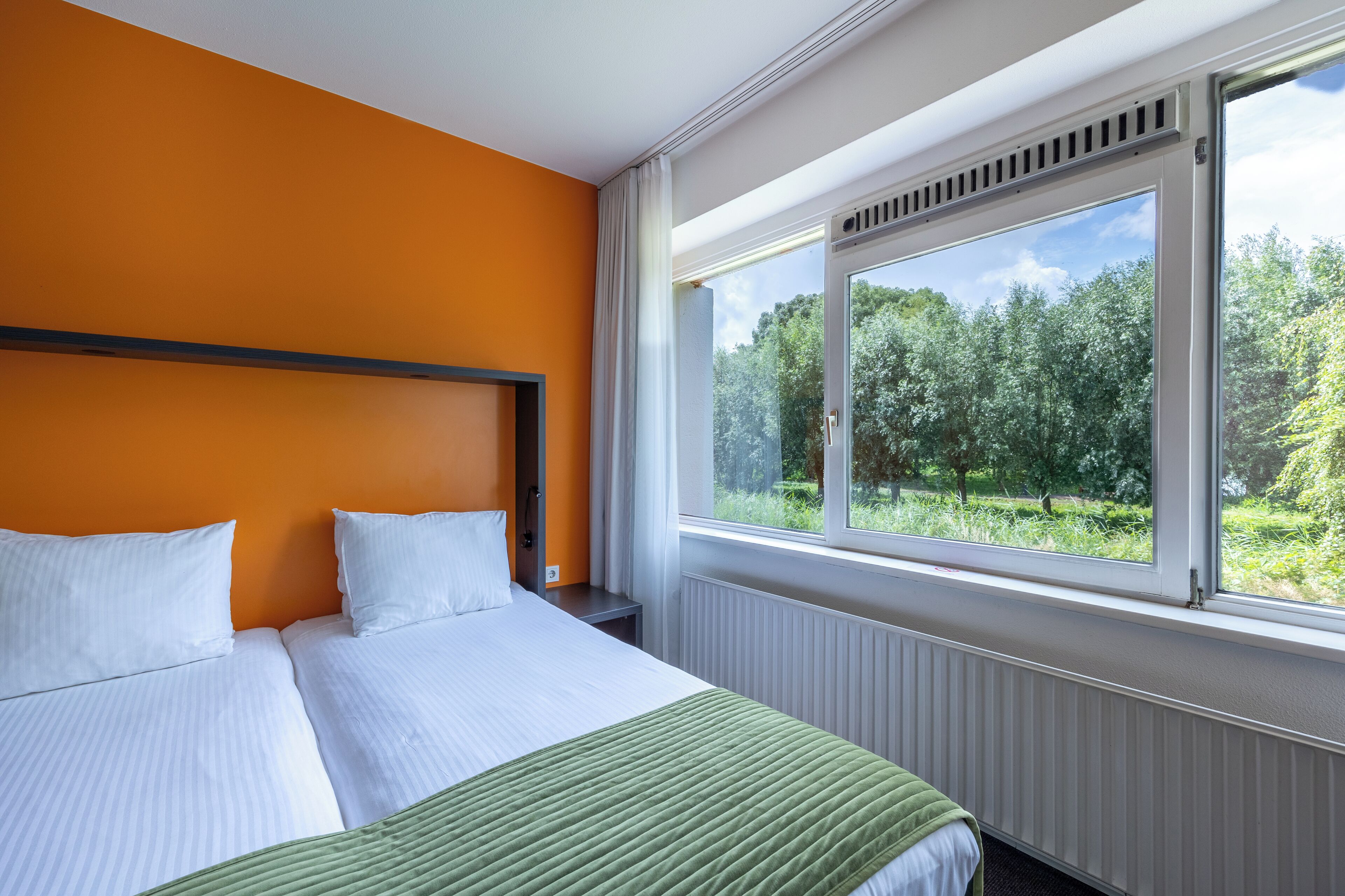 Habitación de hotel con una cama grande, ropa de cama blanca, una pared naranja y una ventana que da a zonas verdes.
