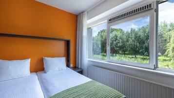 Habitación de hotel con una cama grande, ropa de cama blanca, una pared naranja y una ventana que da a zonas verdes.