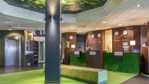 A hotel reception area with a green carpet, a reception desk, clocks displaying different city names, and a check-in/check-out counter.