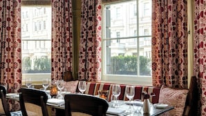 A dining area with a table set for four, a bench, and a view of a building through patterned curtains. - The Gore London – Starhotels Collezione (London)
