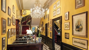 A hotel reception area with a grand staircase, chandeliers, framed artwork, and a reception desk.