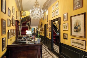 Zona de recepción de un hotel con una escalera monumental, candelabros, obras de arte enmarcadas y un mostrador de recepción.