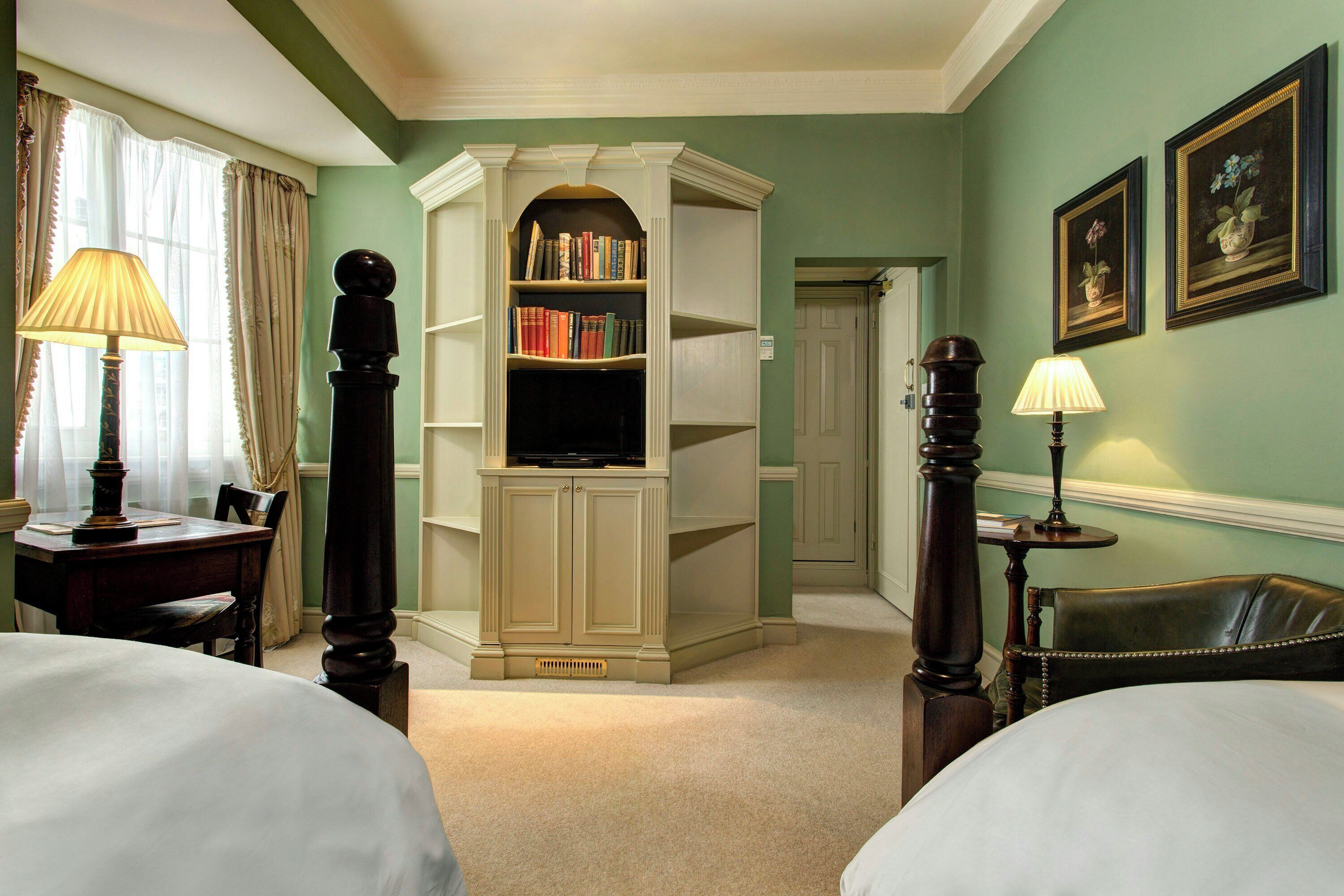 A hotel room with a large bookshelf, two lamps, a desk, and a chair.