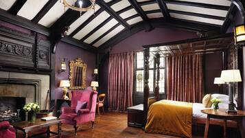A room with a fireplace, a bed with a yellow bedspread, a pink armchair, and a wooden table with a lamp.