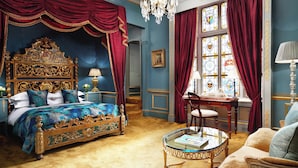 A luxurious bedroom with a large ornate bed, a glass coffee table, and a view of a cityscape through a window with stained glass.