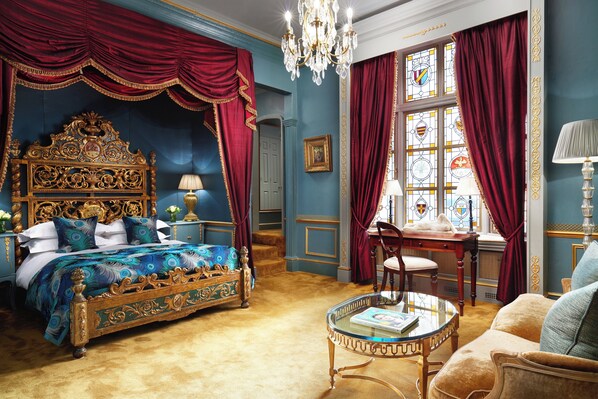 A luxurious bedroom with a large ornate bed, a glass coffee table, and a view of a cityscape through a window with stained glass. - The Gore London – Starhotels Collezione (London)