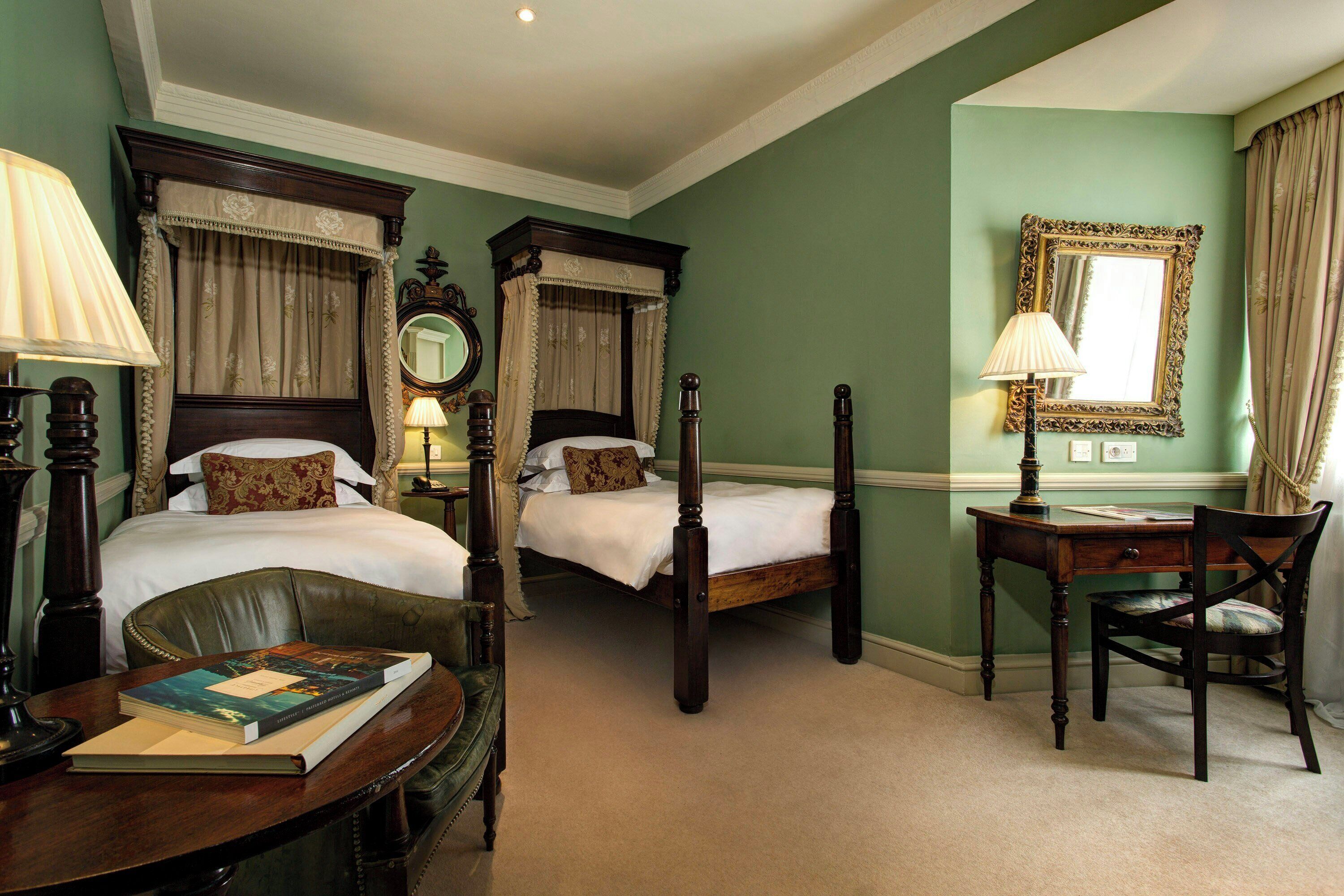 A hotel room with two single beds, a desk with a chair, a lamp, a mirror, and a book on the desk.