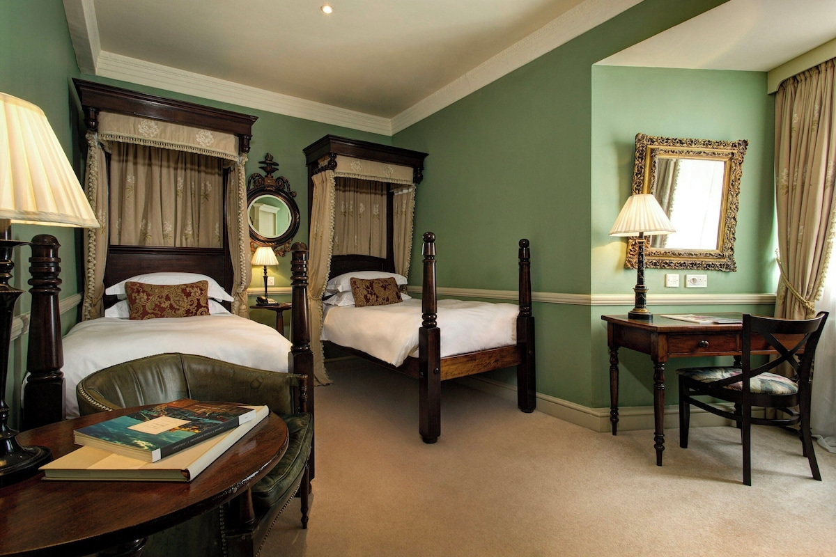 A hotel room with two single beds, a desk with a chair, a lamp, a mirror, and a book on the desk.