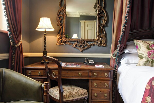 A room with a wooden desk, a chair, a bed with a floral pillow, a mirror, and a lamp.