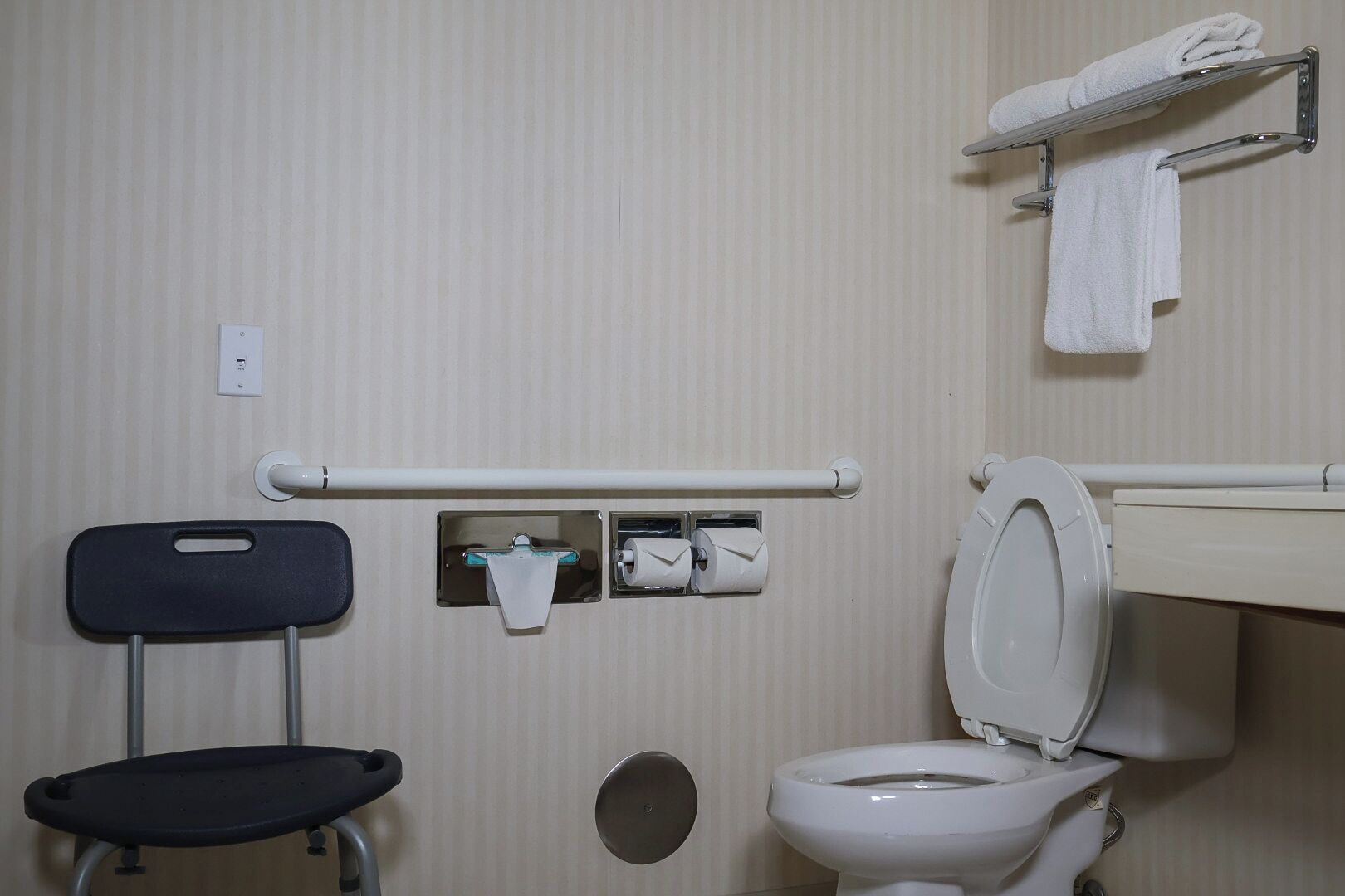 A standard bathroom with a toilet, a handrail, a towel rack, and a chair.