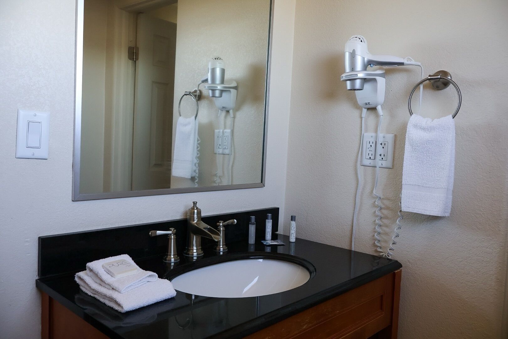 Une salle de bain avec un plan de toilette noir, un lavabo blanc, un miroir, un sèche-cheveux et un porte-serviettes.