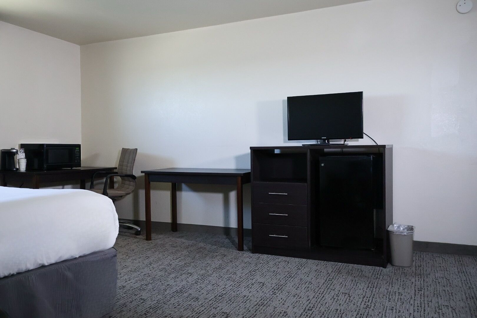 Quarto de hotel com cama, escrivaninha com computador e monitor, cadeira e lixeira.