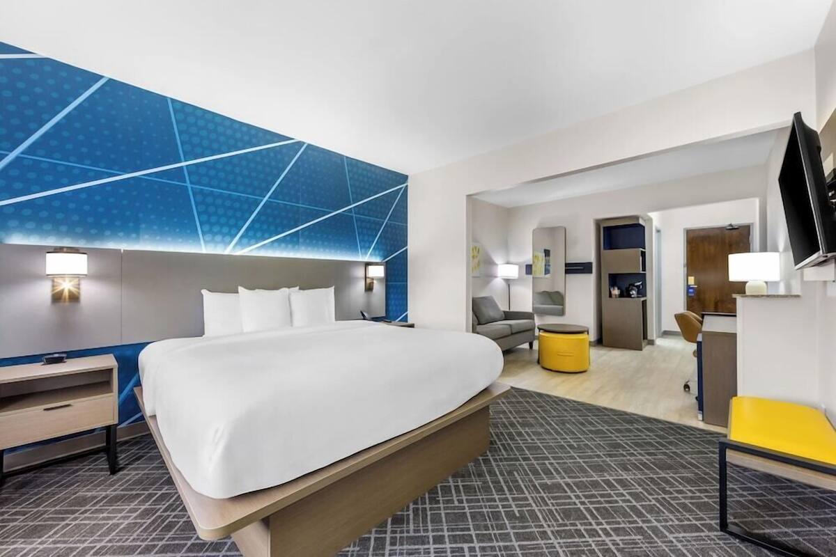 A modern hotel room with a large bed, a desk, a chair, a TV, and a bathroom visible through an open door.