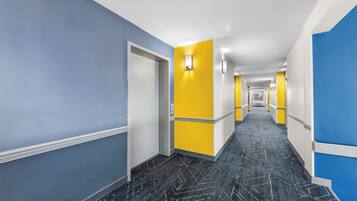 A modern hotel corridor with a geometric patterned carpet, blue and yellow walls, and a long hallway leading to a door.