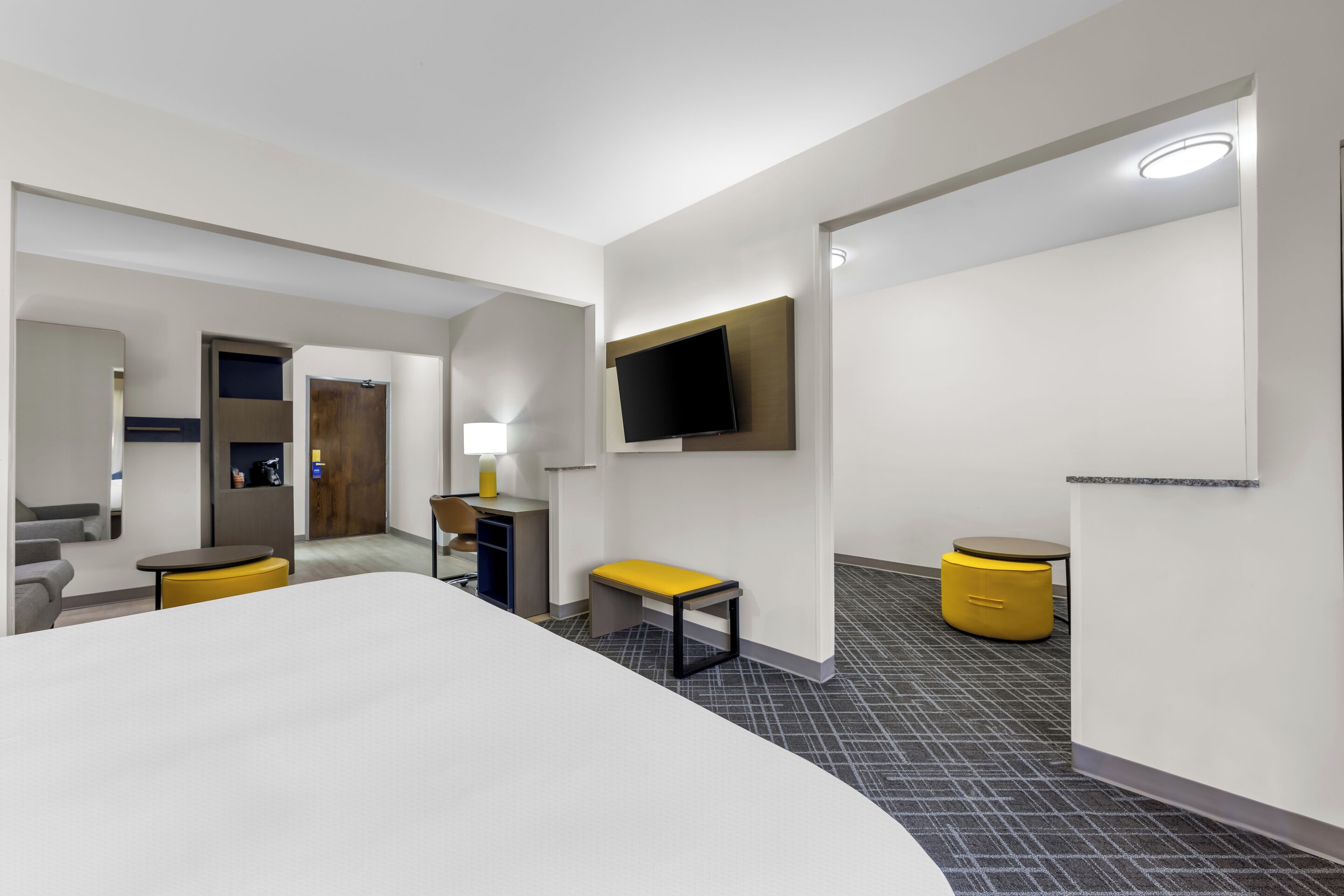 A modern hotel room with a large bed, a desk, a chair, a TV mounted on the wall, and a small table with a bench.