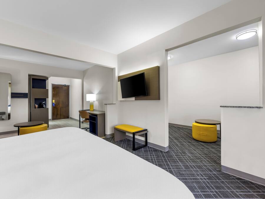 A modern hotel room with a large bed, a desk, a chair, a TV mounted on the wall, and a small table with a bench.