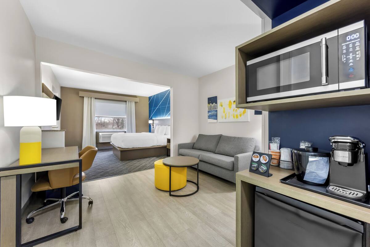 A hotel room with a bed, a sofa, a desk with a chair, a coffee machine, and a microwave.