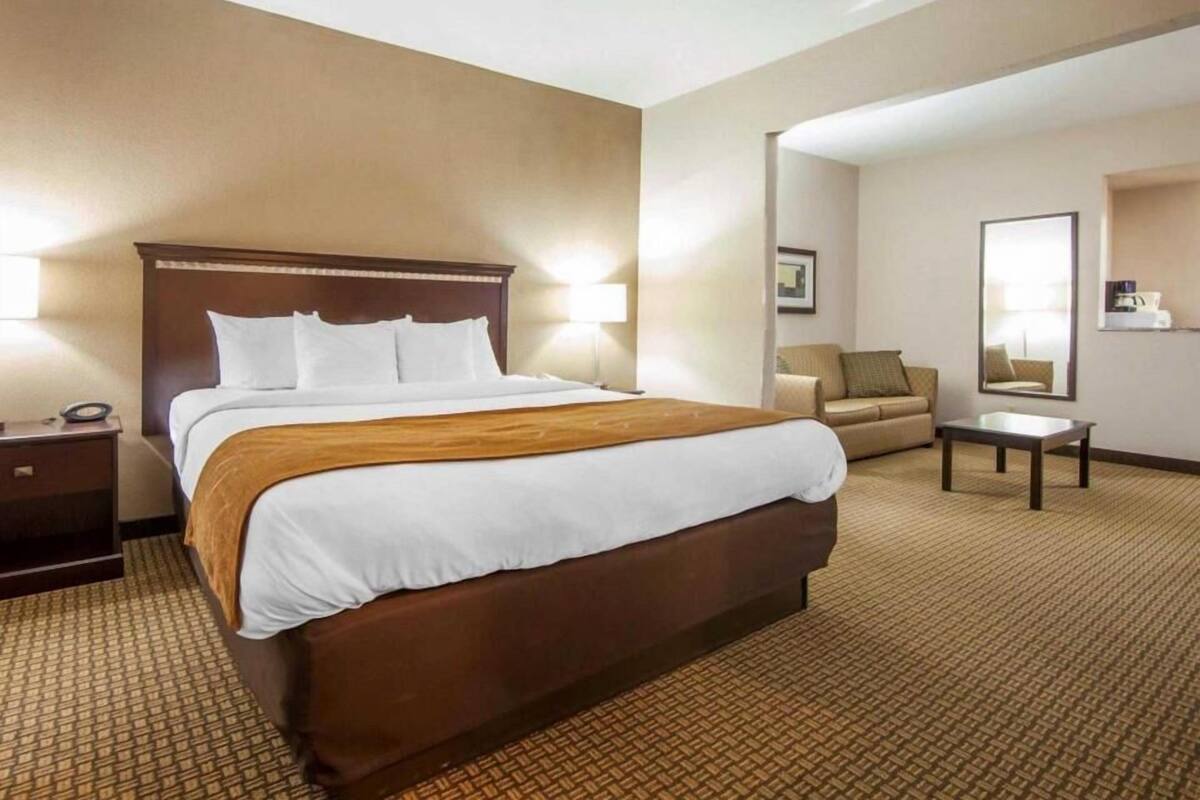 A hotel room with a large bed, a bedside table, a chair, a small table, and a mirror.