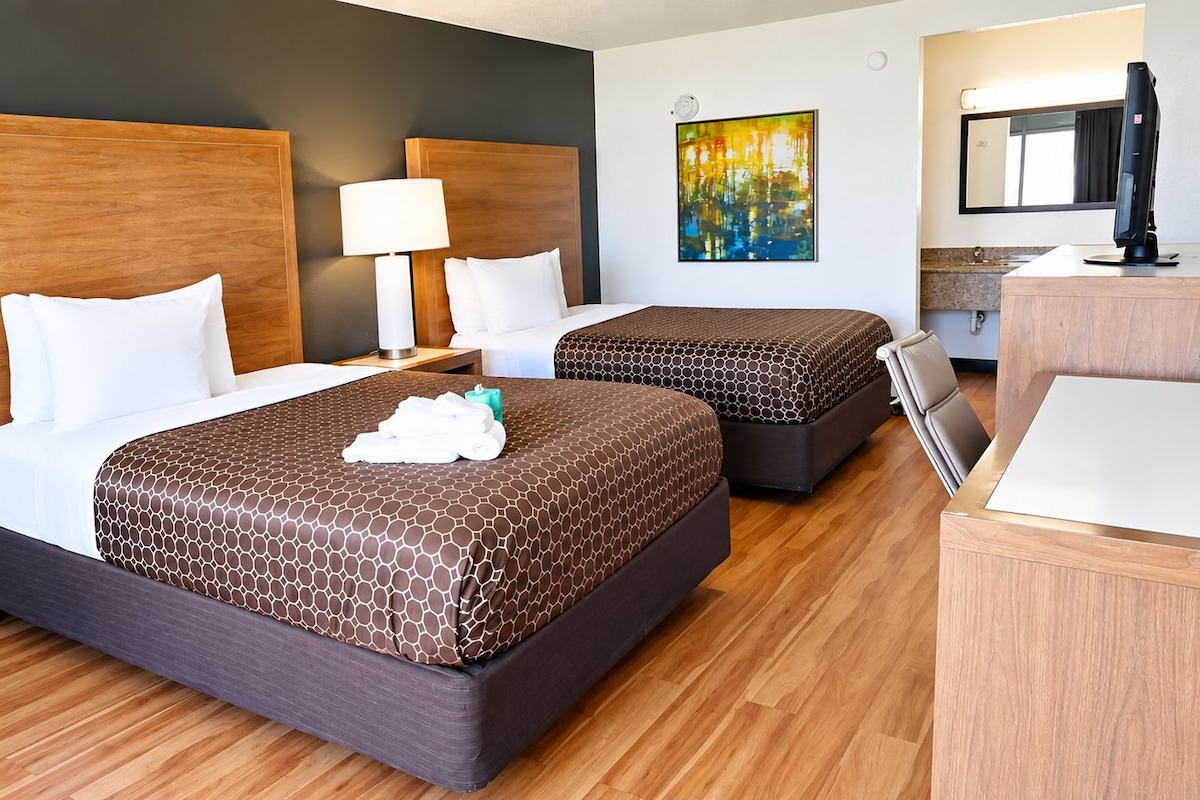 A hotel room with two beds, a desk, a chair, a lamp, and artwork on the wall.