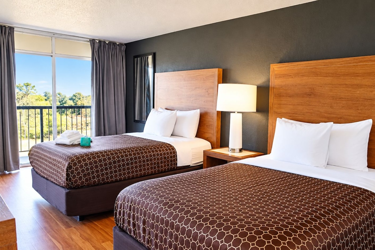 A hotel room with two beds, a nightstand, a lamp, and a view of the outdoors.
