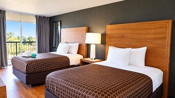 A hotel room with two beds, a nightstand, a lamp, and a view of the outdoors.