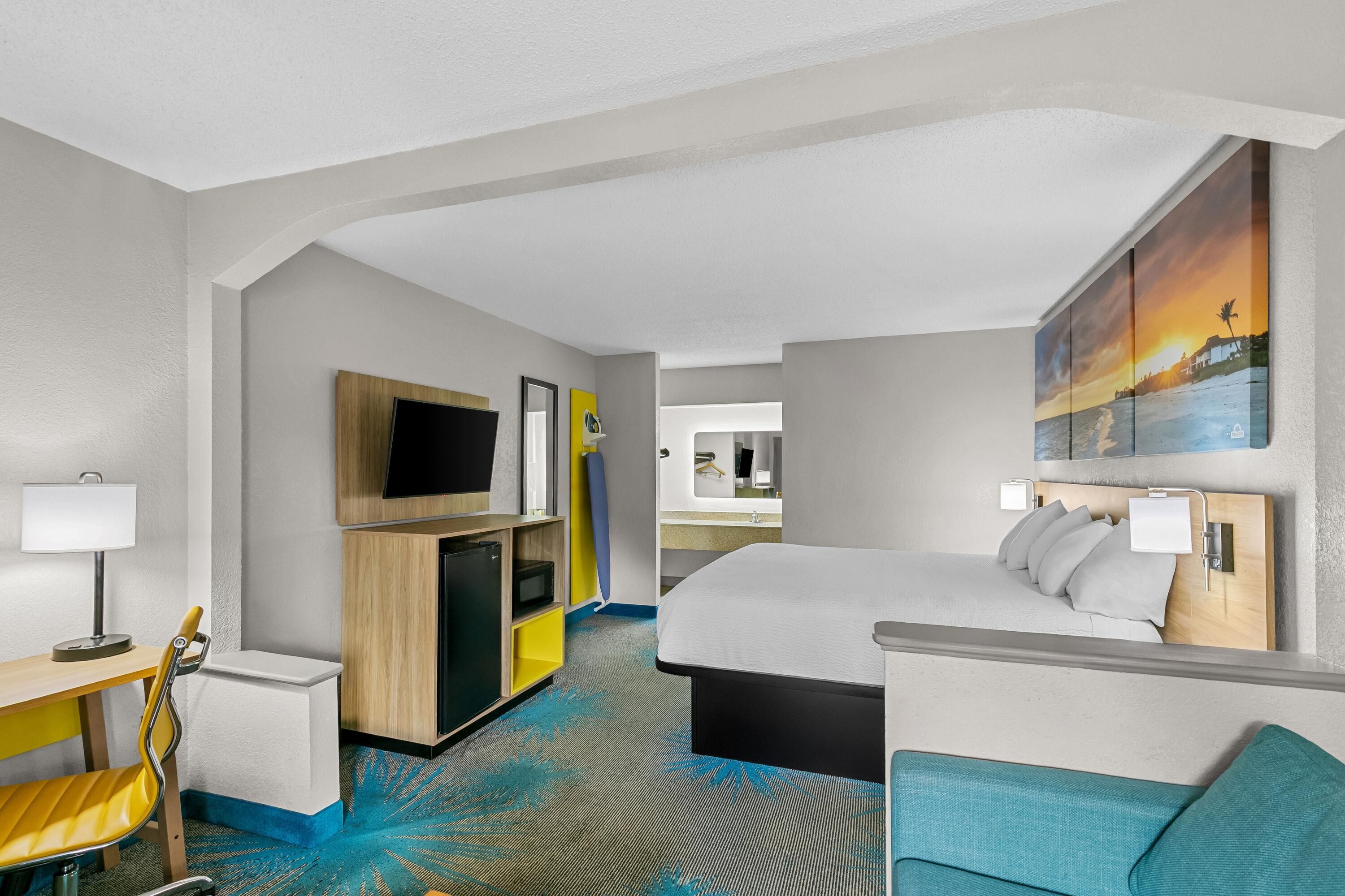 A hotel room with a bed, a desk with a lamp, a TV, a mirror, and a painting on the wall.