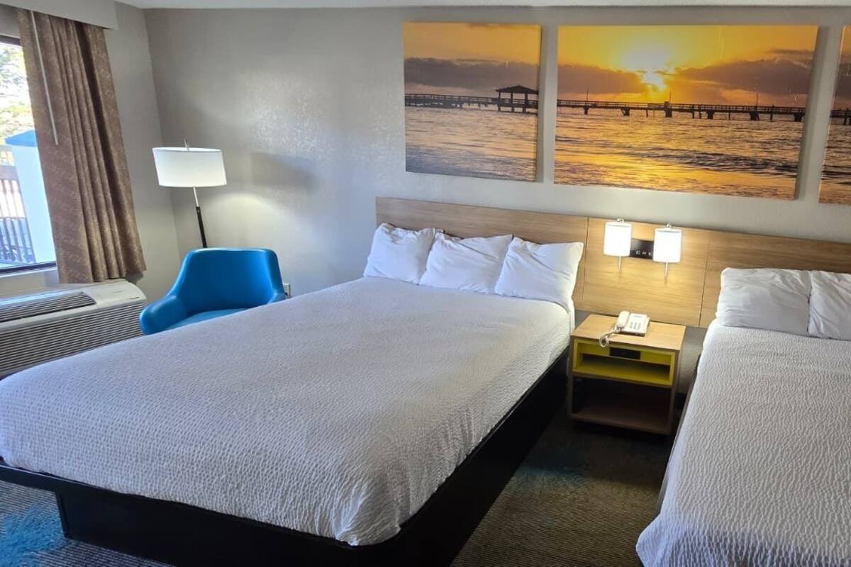 A hotel room with two beds, a desk, a chair, and a wall with sunset paintings.