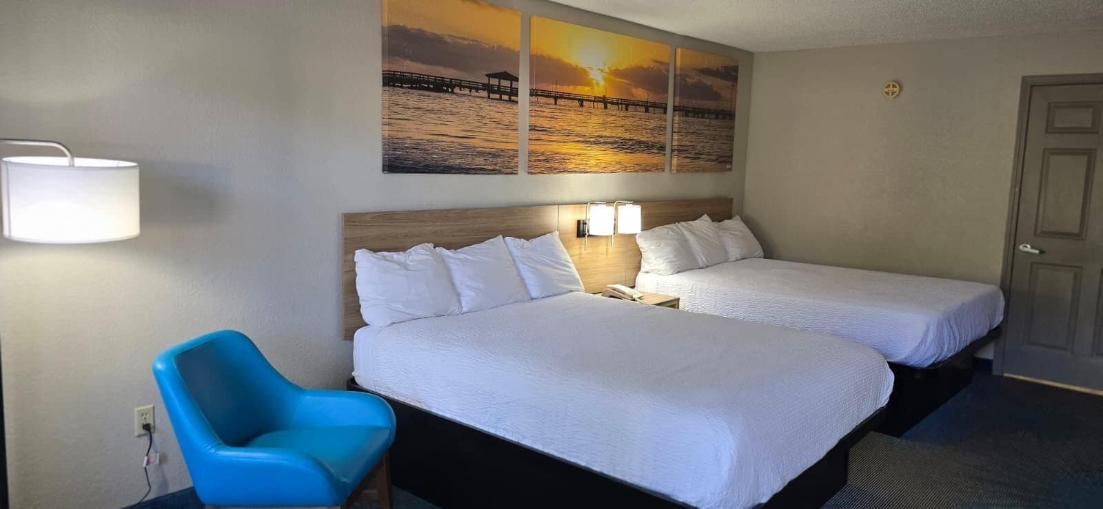 Una habitación de hotel con dos camas, una silla azul y una pared con pinturas de atardecer.