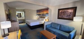 A hotel room with a bed, a blue sofa, a wooden coffee table, a desk with a lamp, and a painting on the wall. - Days Inn by Wyndham Elberton (Elberton)