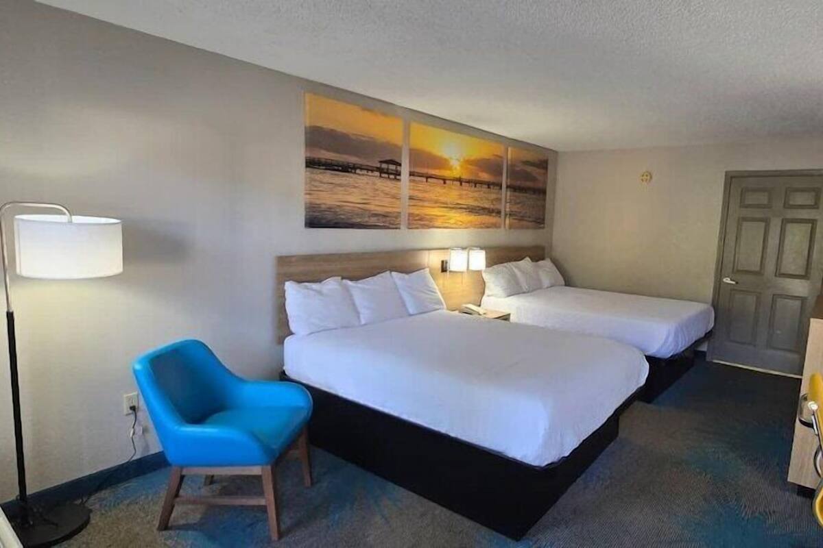 A hotel room with two beds, a desk, a chair, a lamp, and a painting of a sunset.